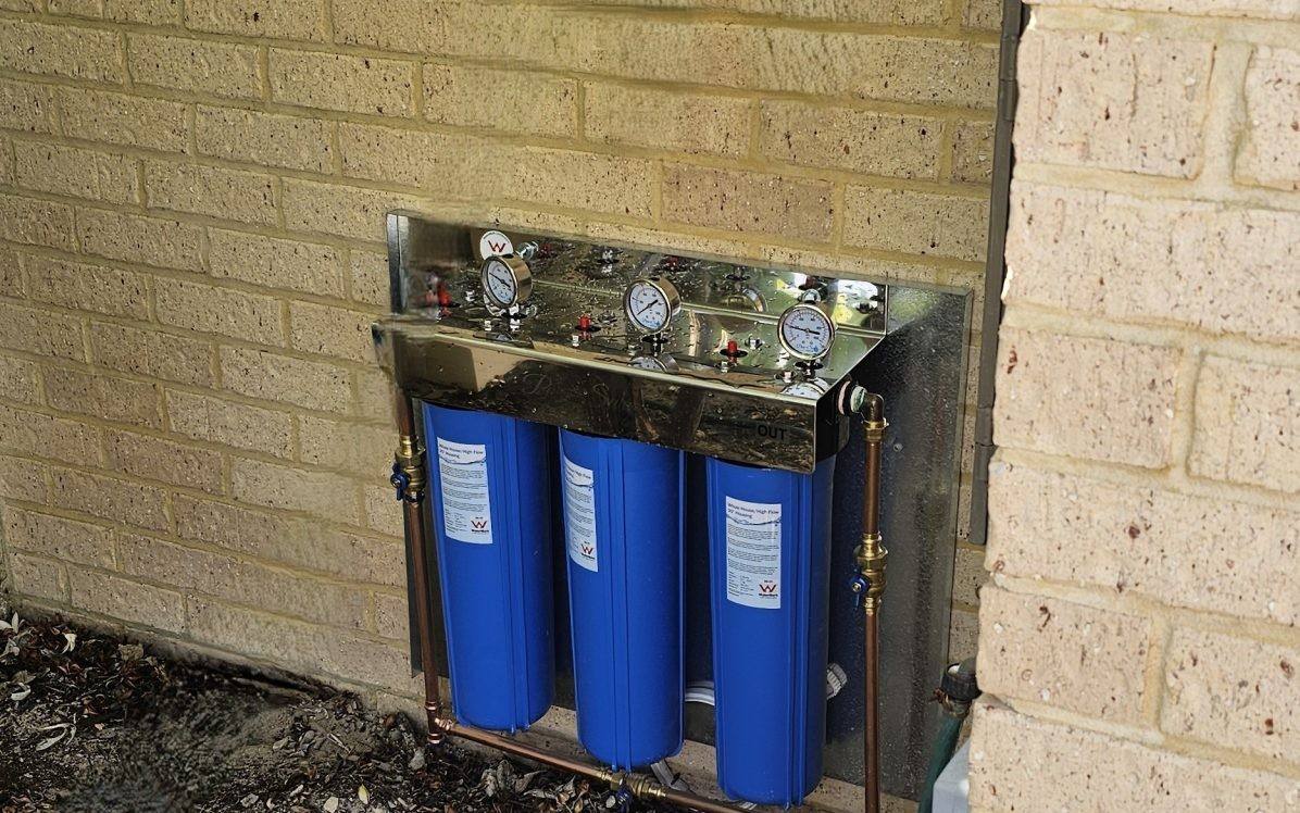 A three-stage whole-house filter with blue housings on a limestone-brick wall beside a frangipani