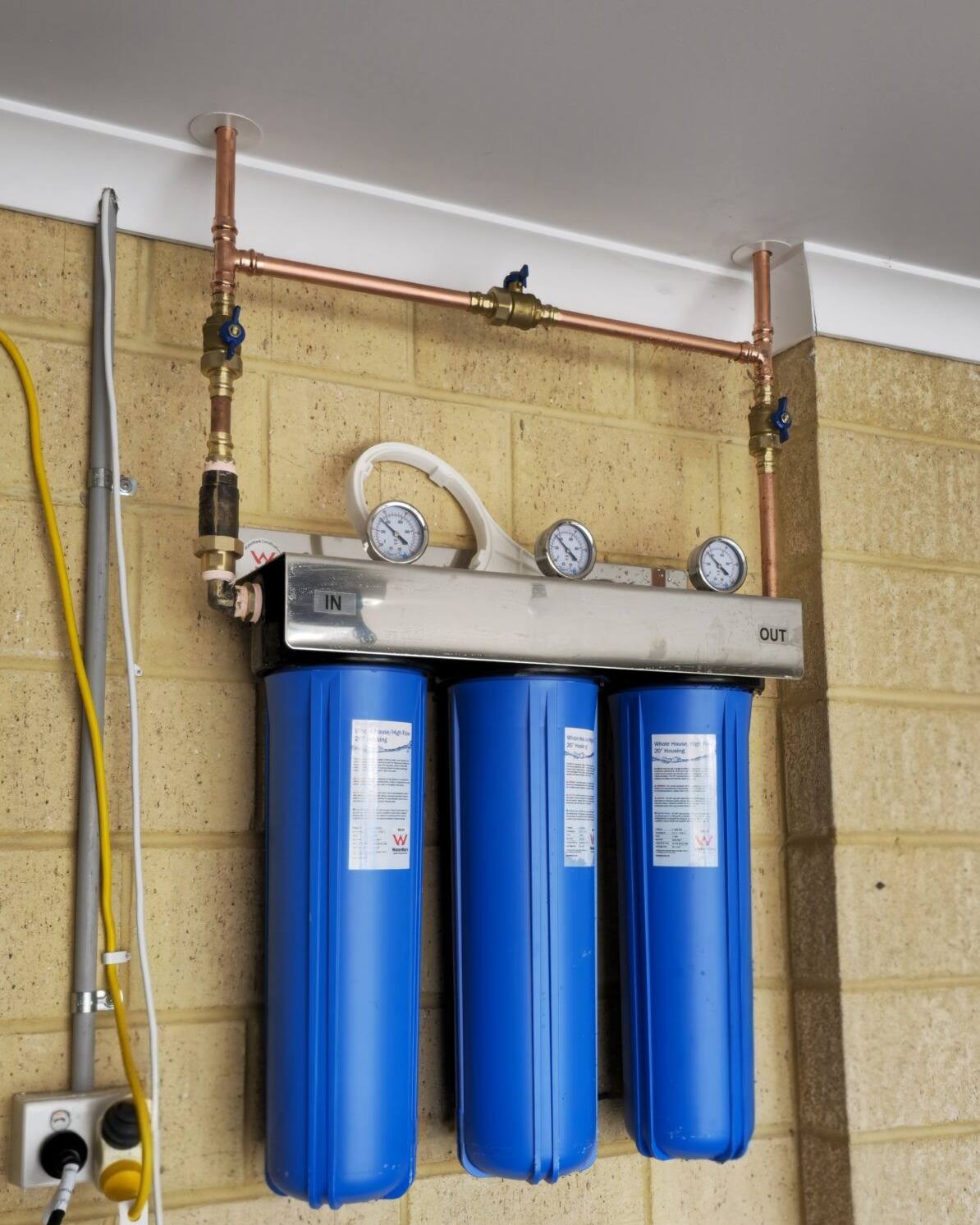 Three large blue filter housings on a stainless manifold with copper pipe, mounted on a garage wall