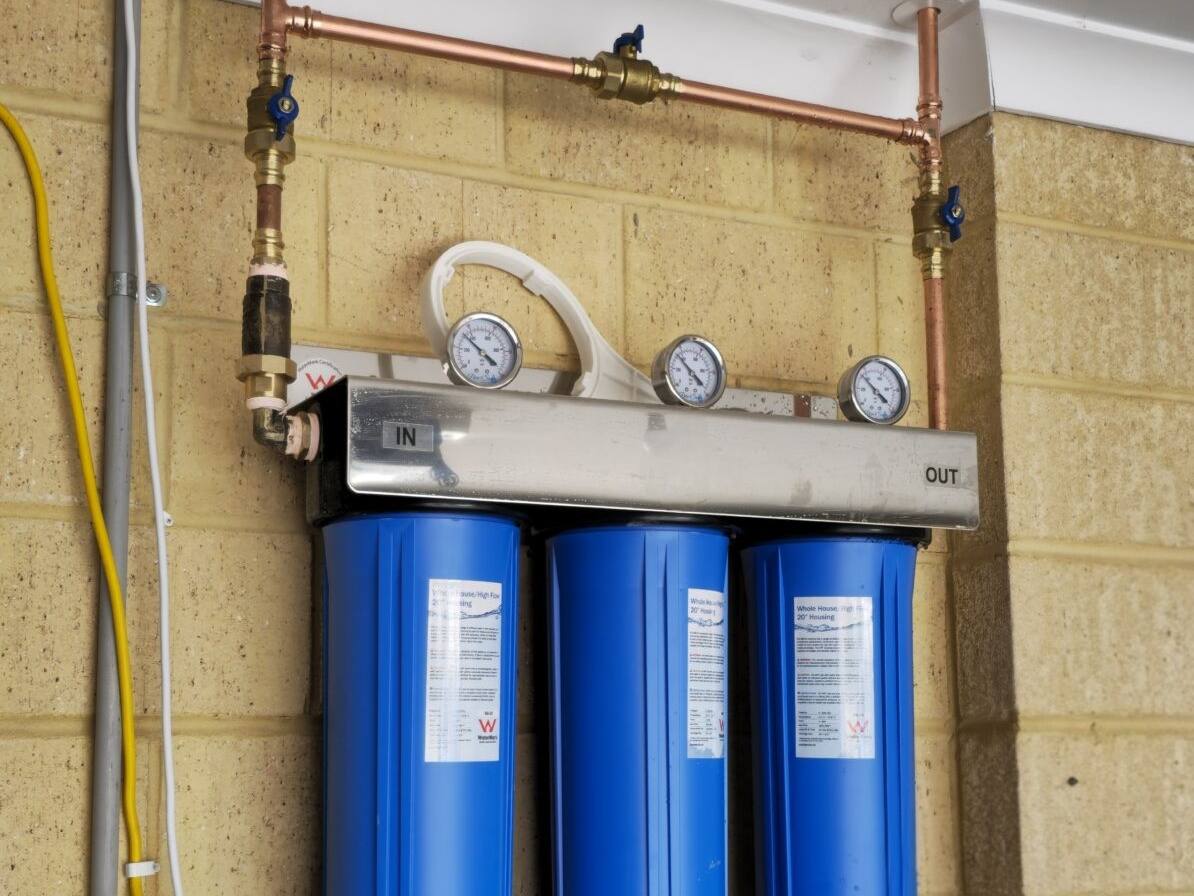 Three large blue filter housings on a stainless manifold with copper pipe, mounted on a garage wall