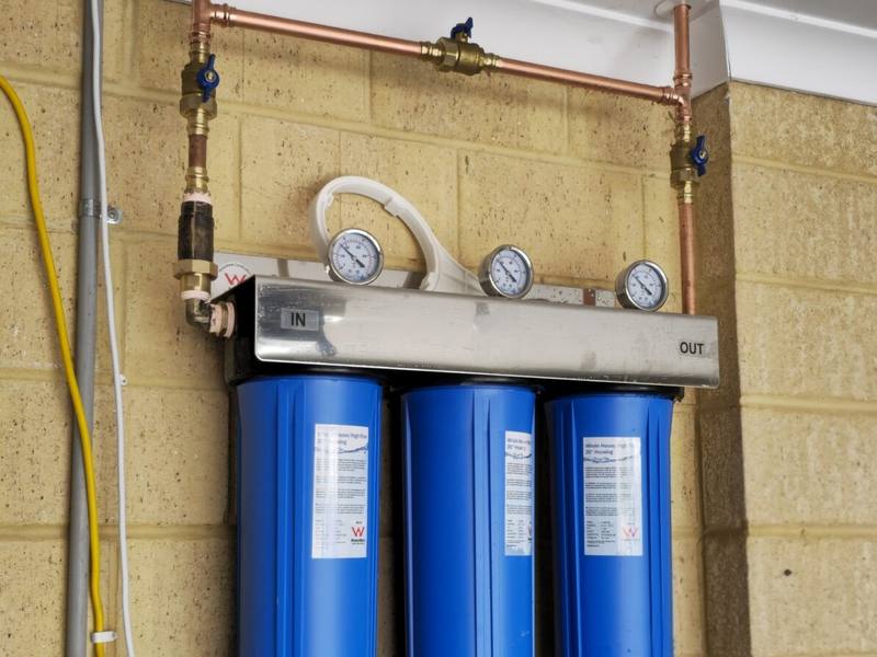 Three large blue filter housings on a stainless manifold with copper pipe, mounted on a garage wall