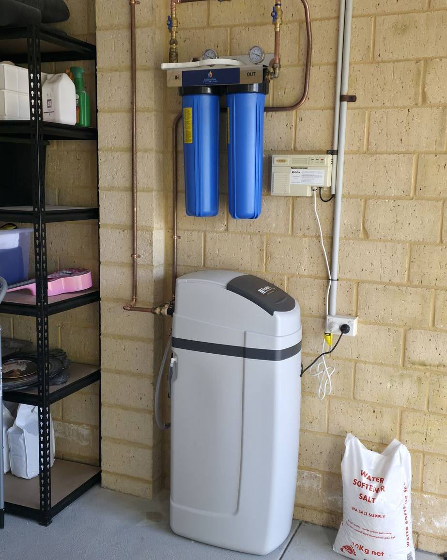 A water softener below two blue filter housings, with a bag of softener salt alongside