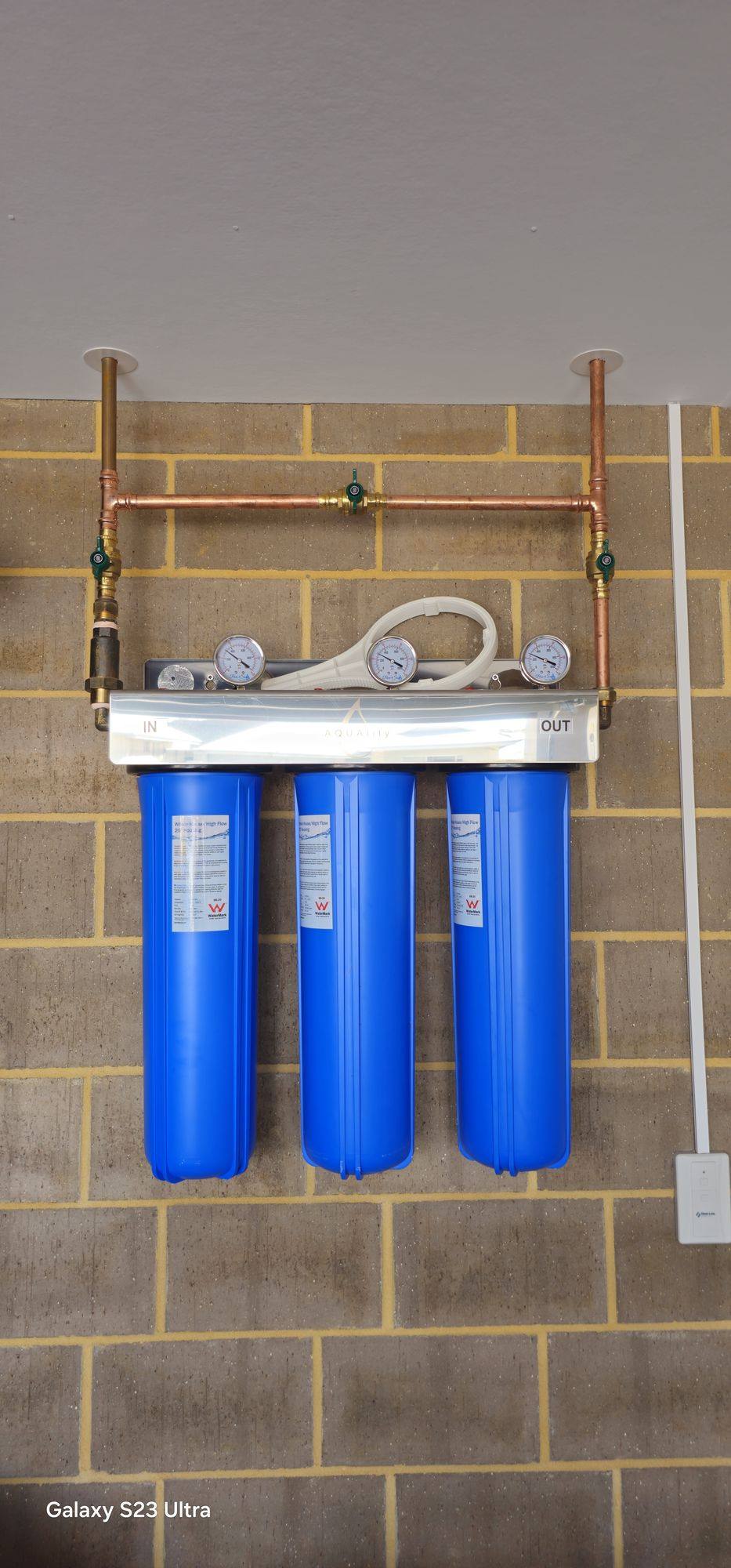 Three blue filter housings with pressure gauges and copper pipework on a brick garage wall