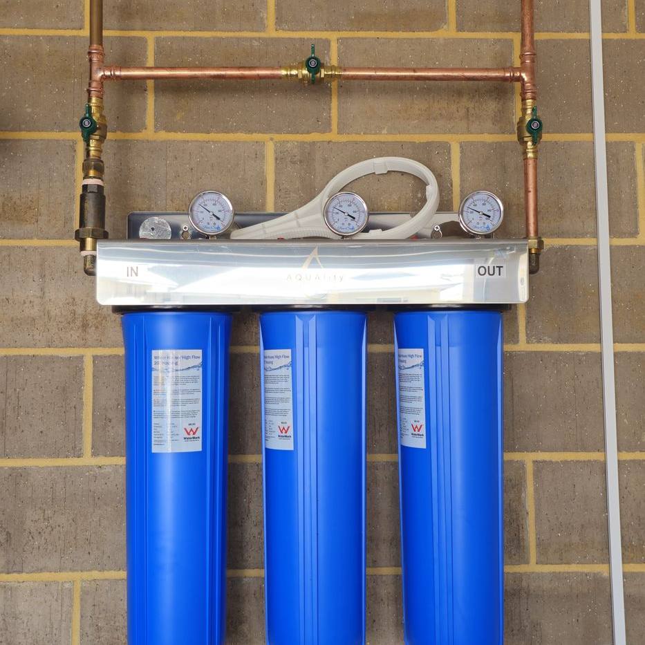 Three blue filter housings with pressure gauges and copper pipework on a brick garage wall